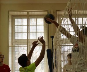 w szkolnej hali gimanstycznej ujęcie na sitakę do gry w siatkówkę, jeden chłopiec skacze i przebija piłkę na drugę połowę natomiast przeciwnik wyrywa się do bloku. W tle zawodnicy przyglądają się zawodnikom z obu drużyn