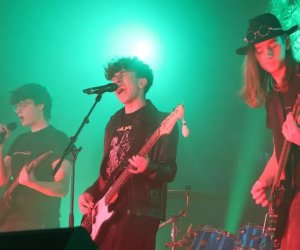 na pierwszym planie na tle zielonych świateł występują trzej mężczyźni grający na gitarach, z których dwóch dodatkowo śpiewa do mikrofonów na statywach. W tle z prawej strony widać perkusistę grającego na niebieskiej perkusji