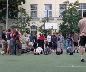 na boisku szkolnym zewnętrznym jest zebrane duże grono uczniów, które przygląda się 5 nastolatkom wykonującym ćwiczenie "deskę". W centrum zdjęcia znajduje się komisja. W tle widnieje budynek szkoły